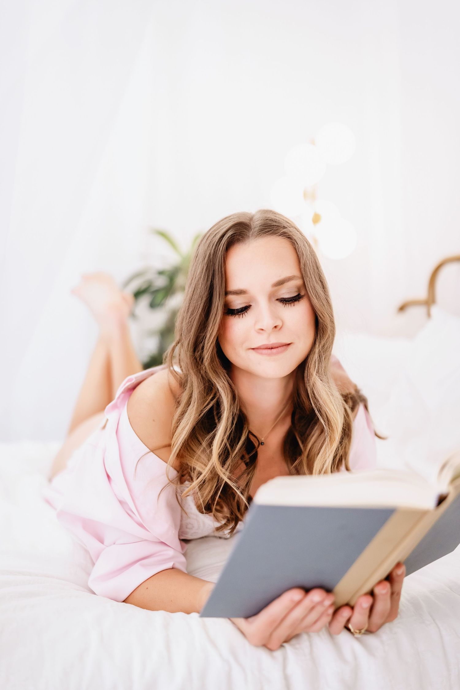 Person in pink loungewear reading a book while lying comfortably on white bedding.