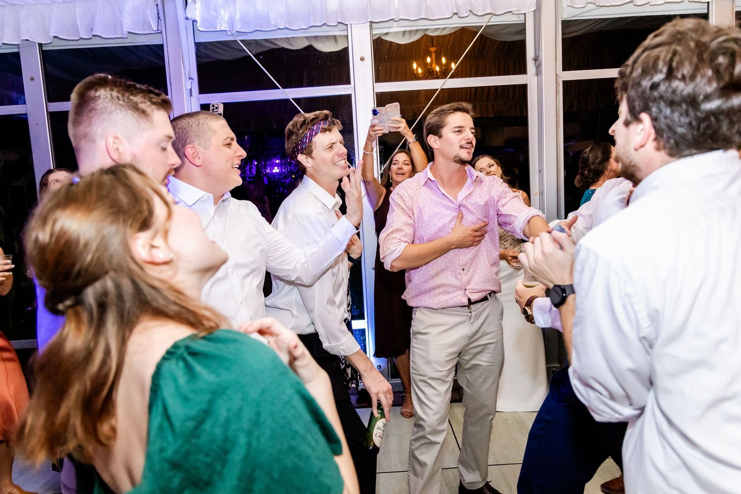 Panoramic view of wedding guests dancing under purple lighting at a tented reception.