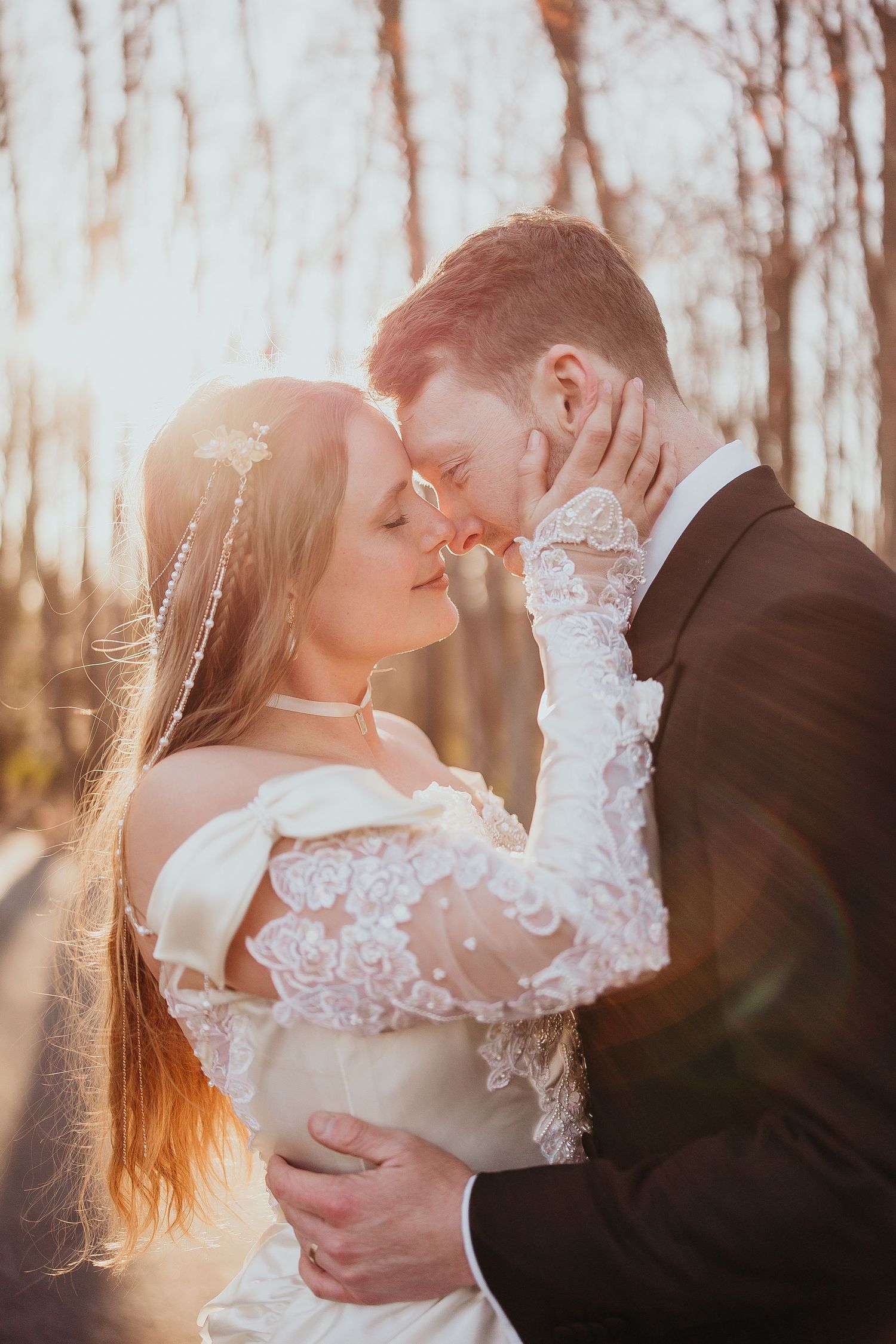 Newlyweds embracing tenderly in golden sunset light, bride in off-shoulder lace gown touching grooms face.