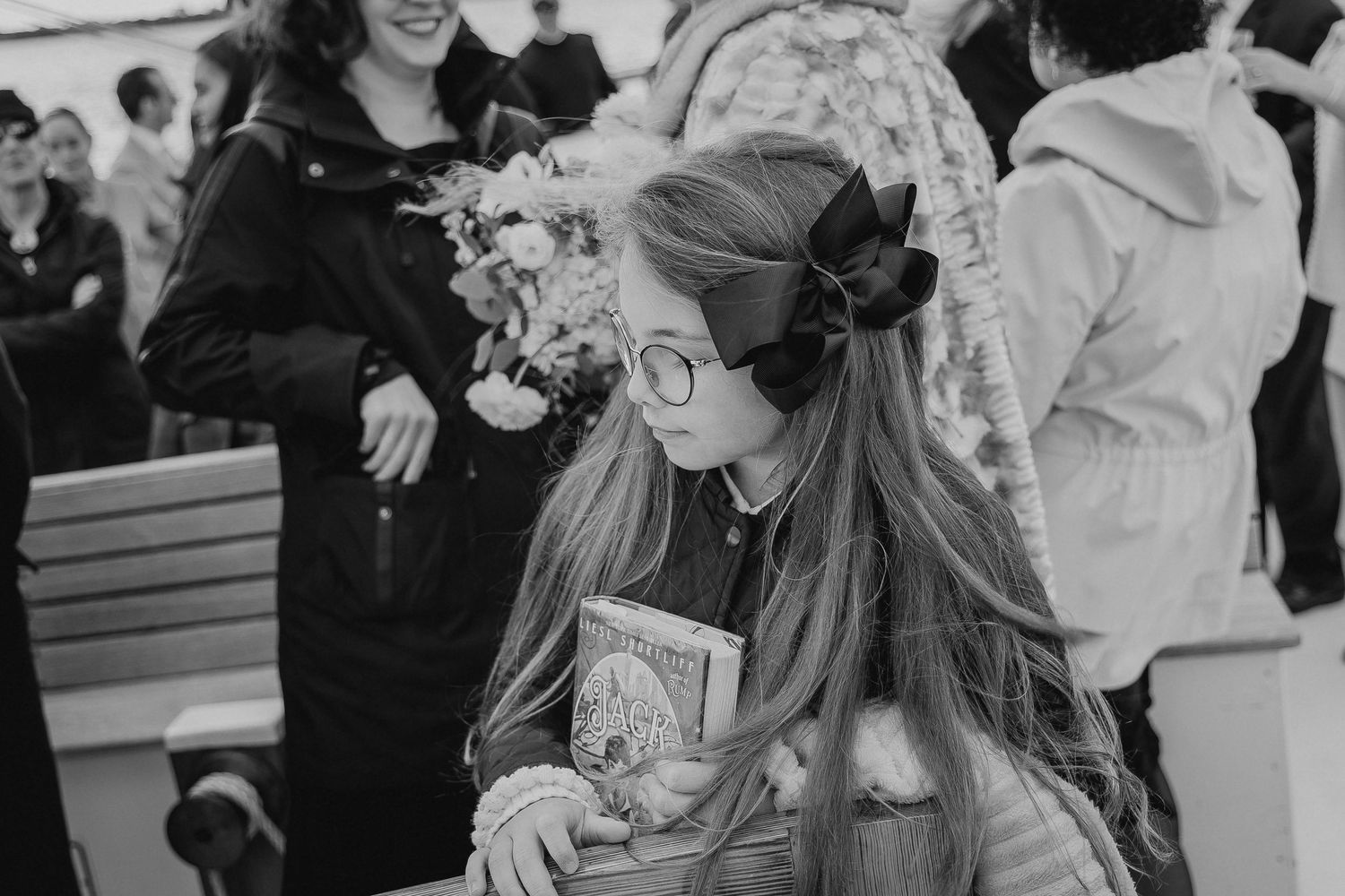 Young girl with long hair and glasses holding a book at an outdoor gathering, black and white photo.