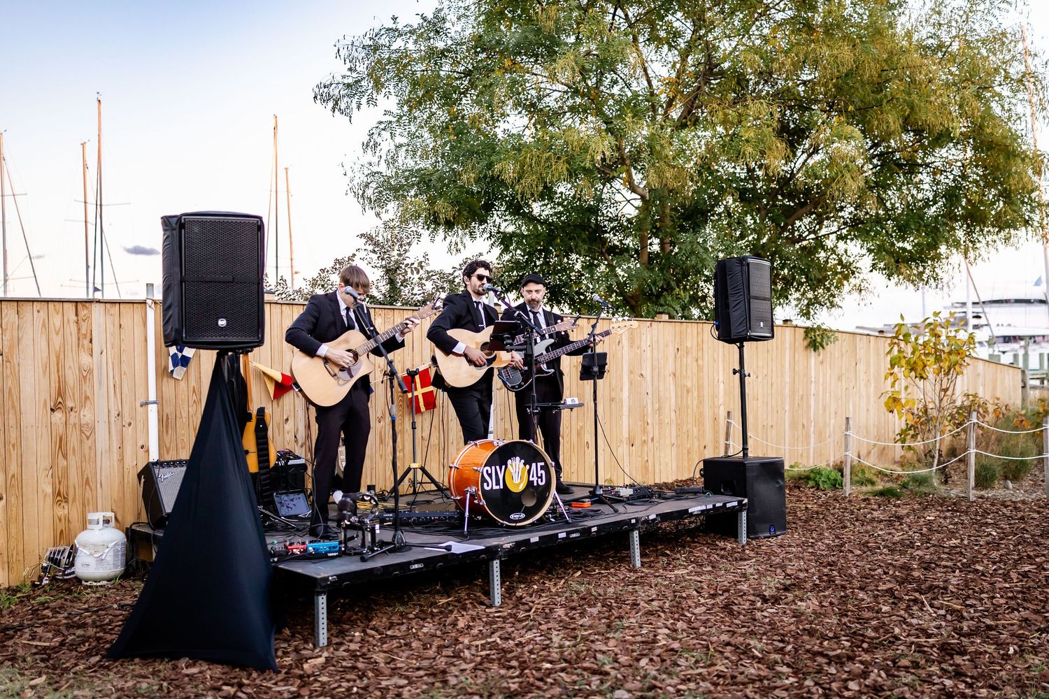 Two musicians performing acoustic guitars on an outdoor stage with speakers and a wooden fence backdrop.