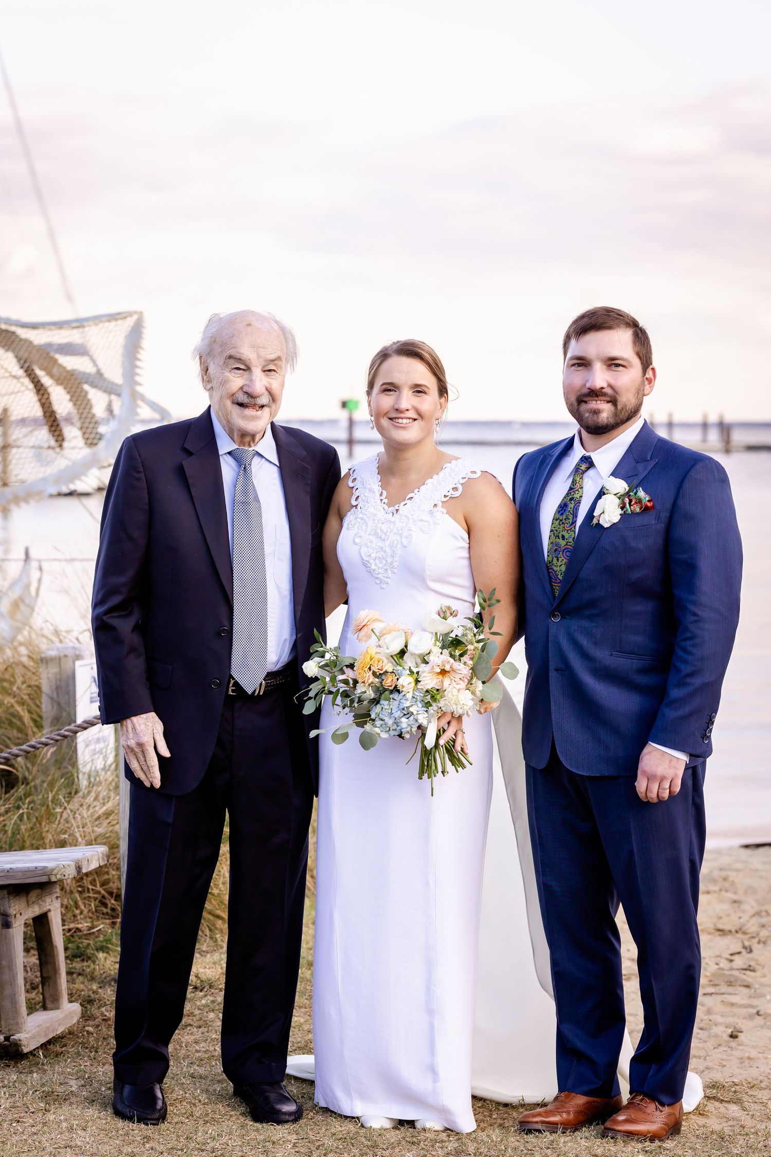 Bride in white gown holds bouquet flanked by two men in suits near waterfront at outdoor wedding.