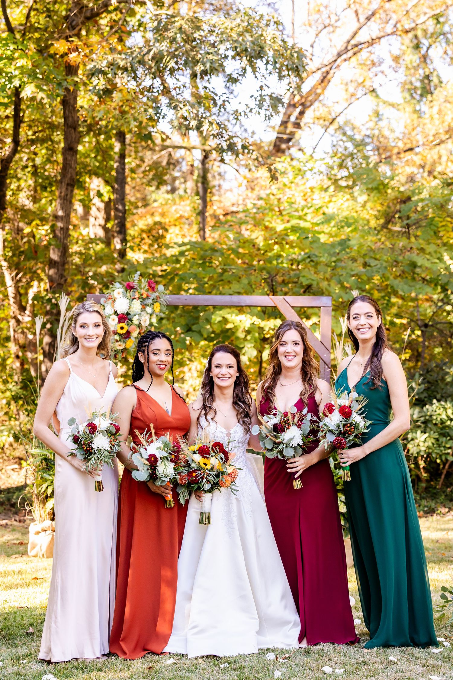 Bridesmaids in burgundy and emerald dresses hold bouquets while standing outdoors.