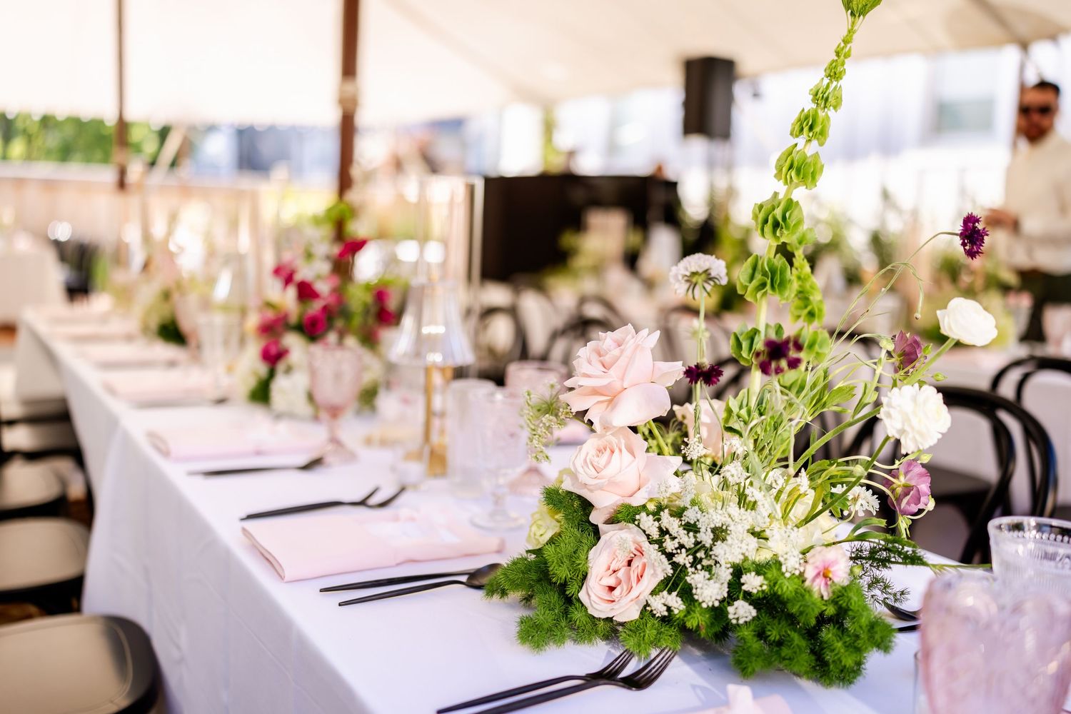 Wedding reception tables decorated with pink floral arrangements and greenery under white tent.