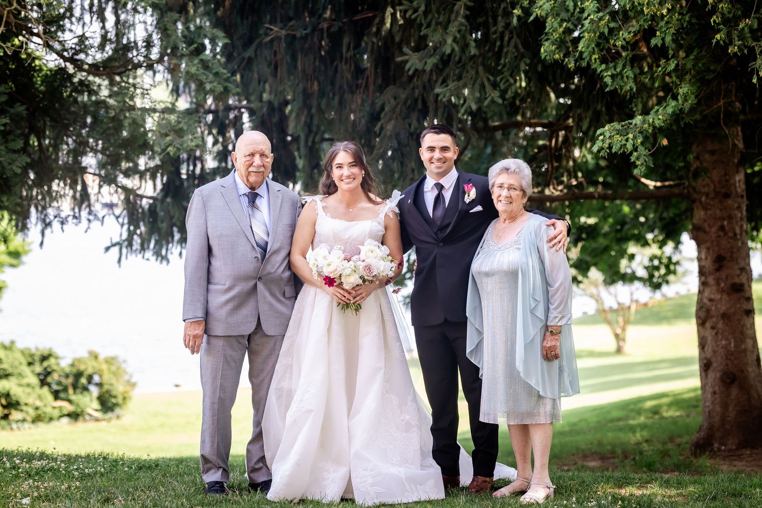 Wedding party poses for formal photos under large evergreen trees outdoors.