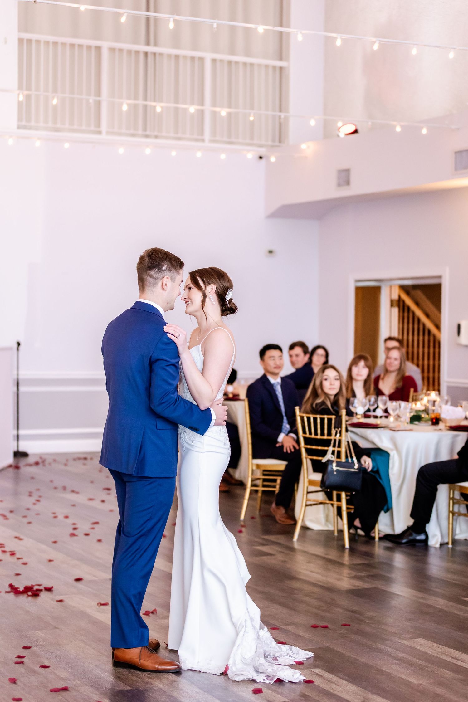 Newlyweds share their first dance in an elegant white reception venue with hardwood floors and rose petals.