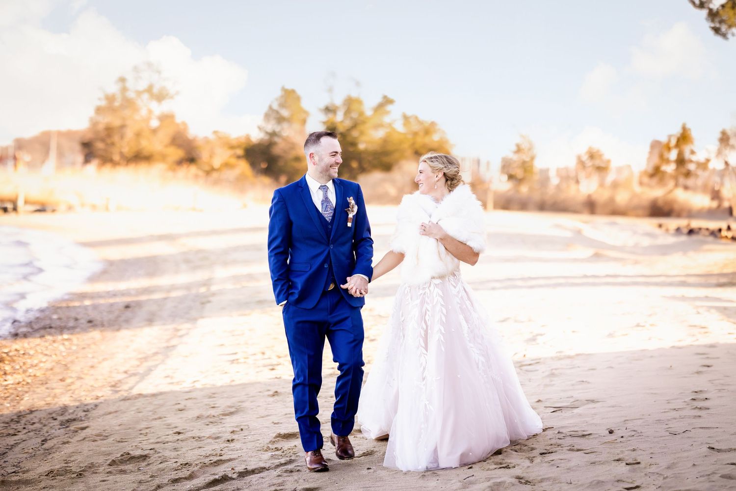 A couple walks together on a sandy path at sunset wearing elegant blue suit and white gown.
