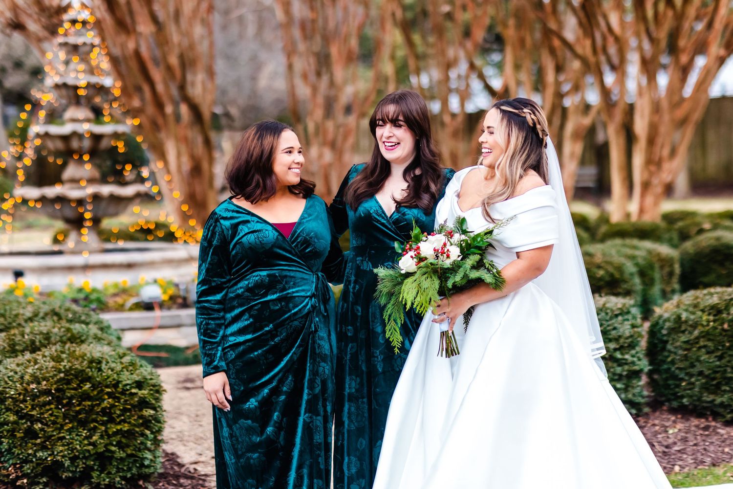 Three women in emerald velvet dresses and white wedding gown laugh together in a garden setting with ornamental bushes.