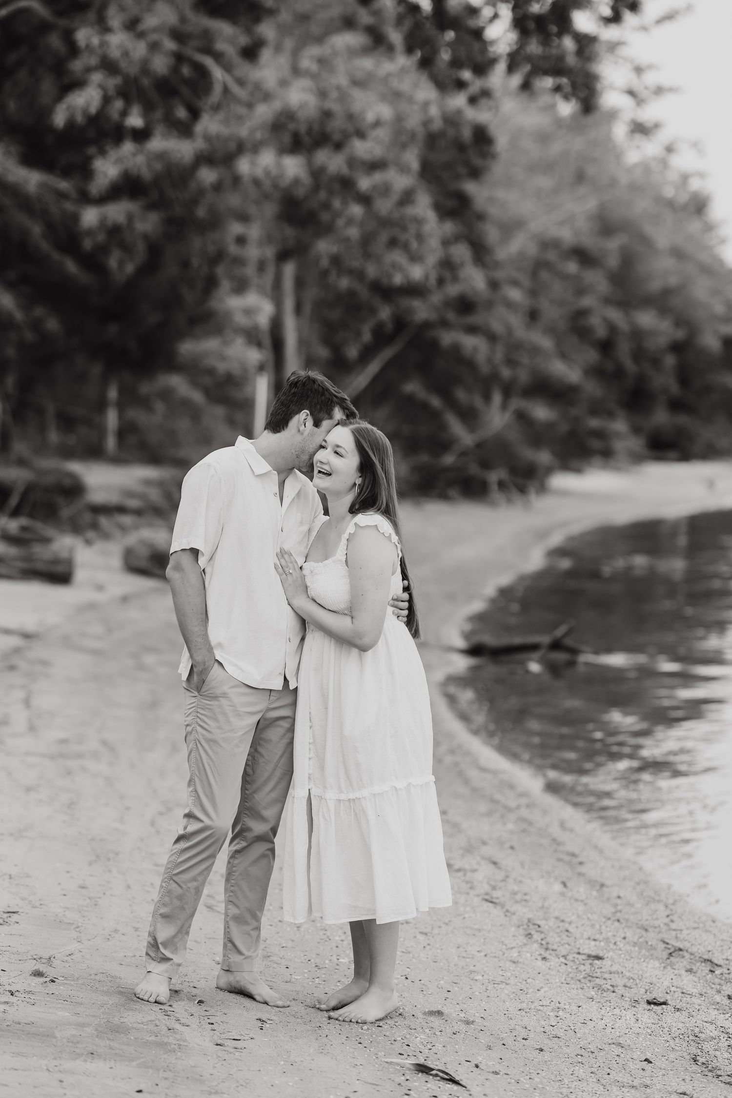 A romantic black and white photograph of a couple sharing an intimate moment on a lakeside beach.