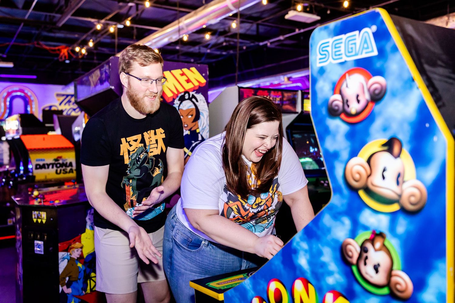 Two people looking down excitedly at an arcade game cabinet featuring colorful Sega monkey characters.