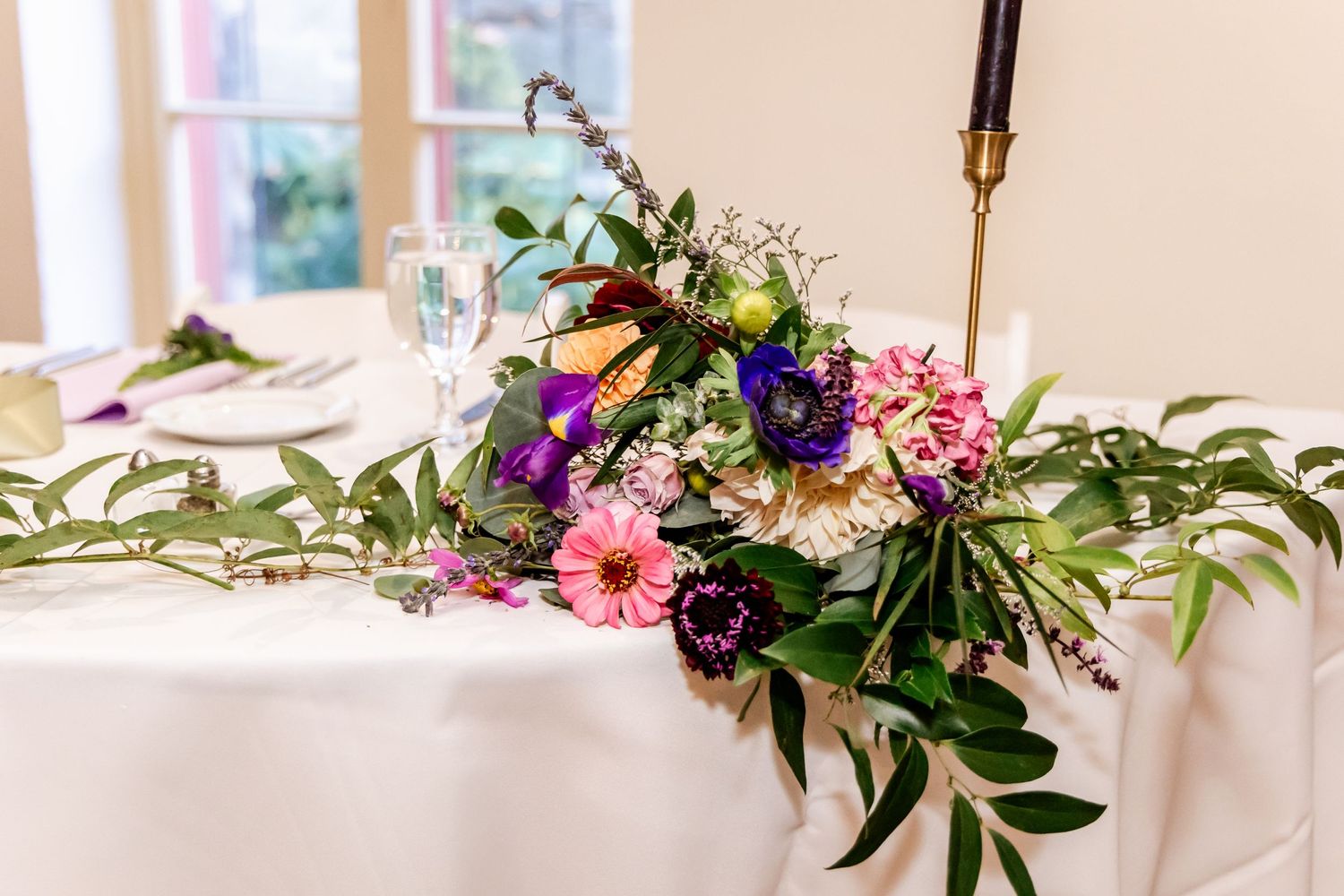 A stunning floral centerpiece with pink and purple blooms adorns a white tablecloth at a wedding reception.