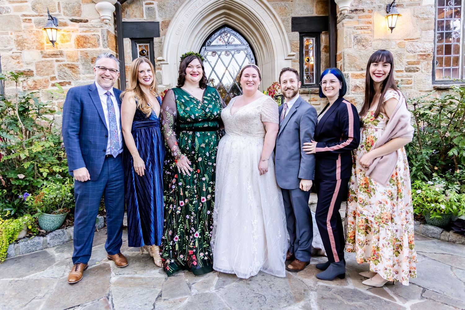 Wedding party poses together outside stone church entrance in colorful formal attire.