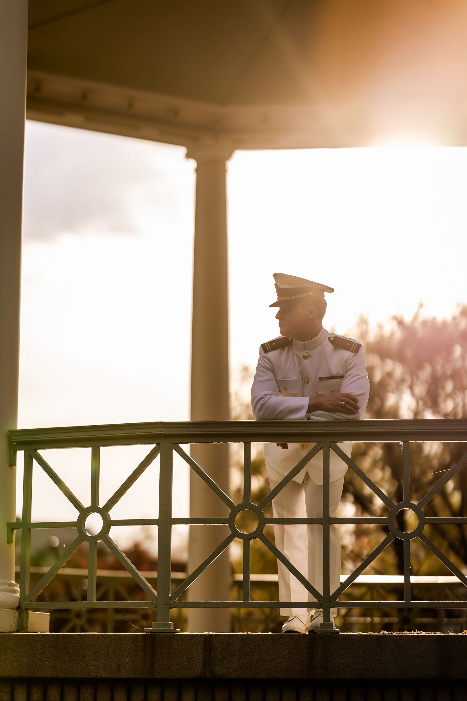 A naval officer in white uniform stands pensively on a porch balcony at sunset with ornate railing details.