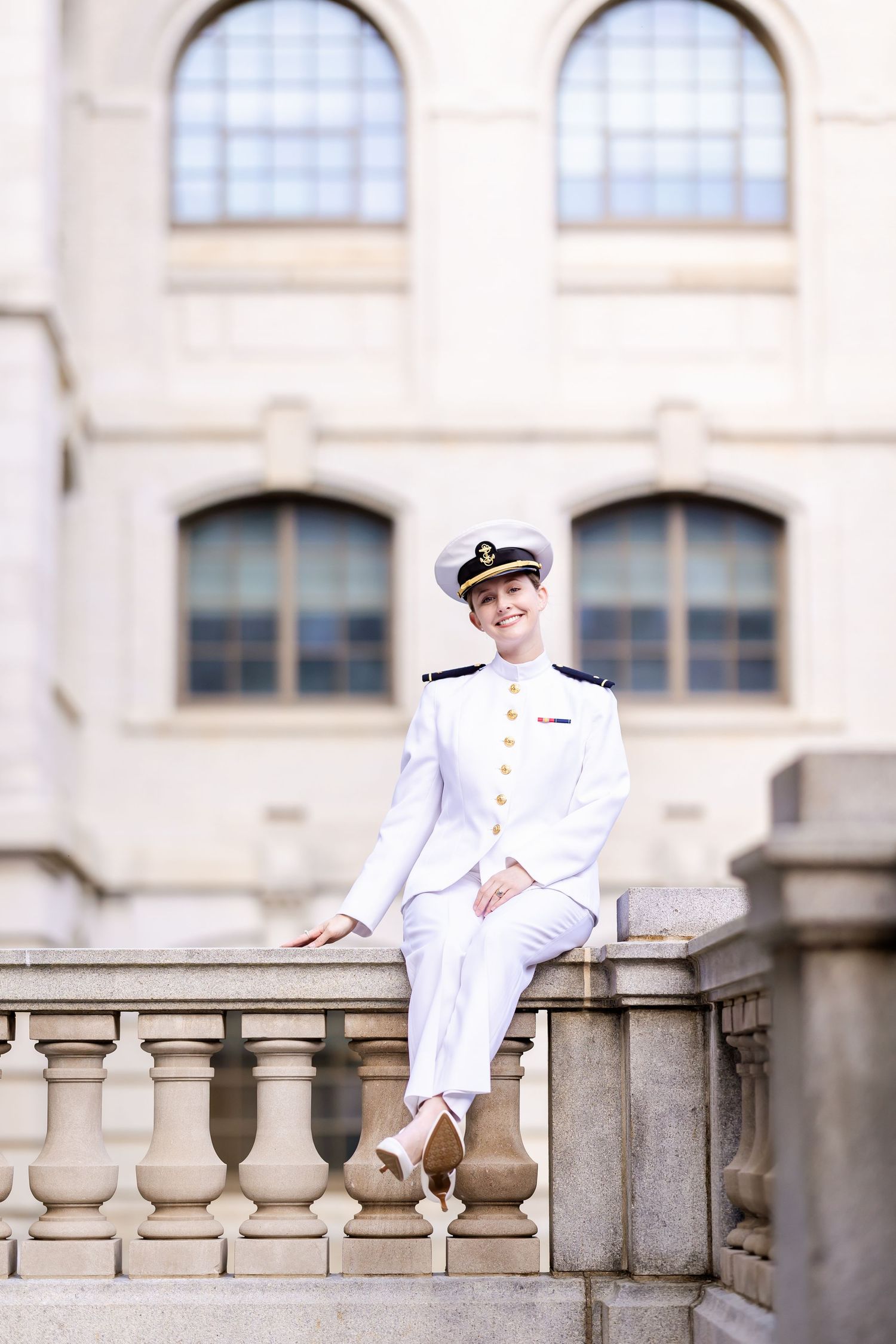 Naval officer in formal white dress uniform sitting on ornate stone balustrade with arched windows in background.