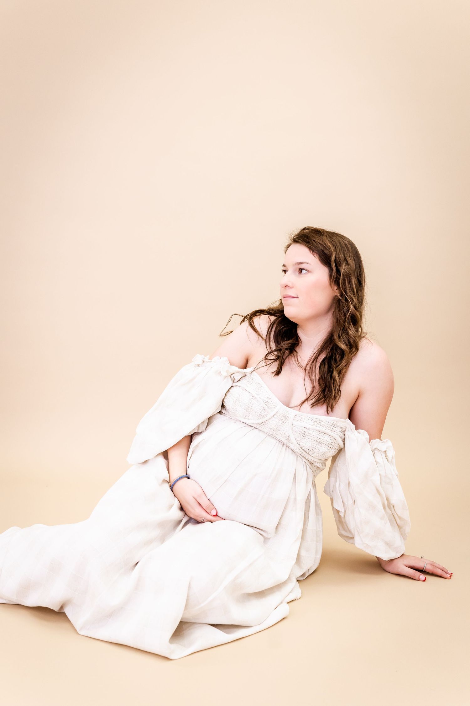 A person in a flowy white off-shoulder dress reclines on a beige background in an elegant studio portrait.