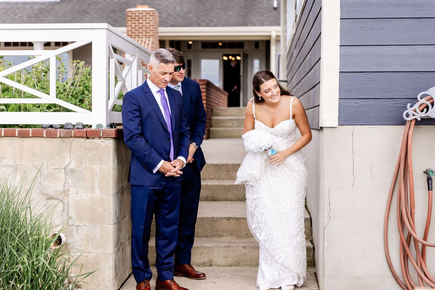 Couple in wedding attire standing on steps outside a gray house.