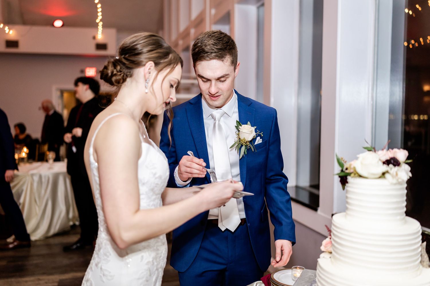 A couple in wedding attire examines their multi-tiered white wedding cake during the reception.