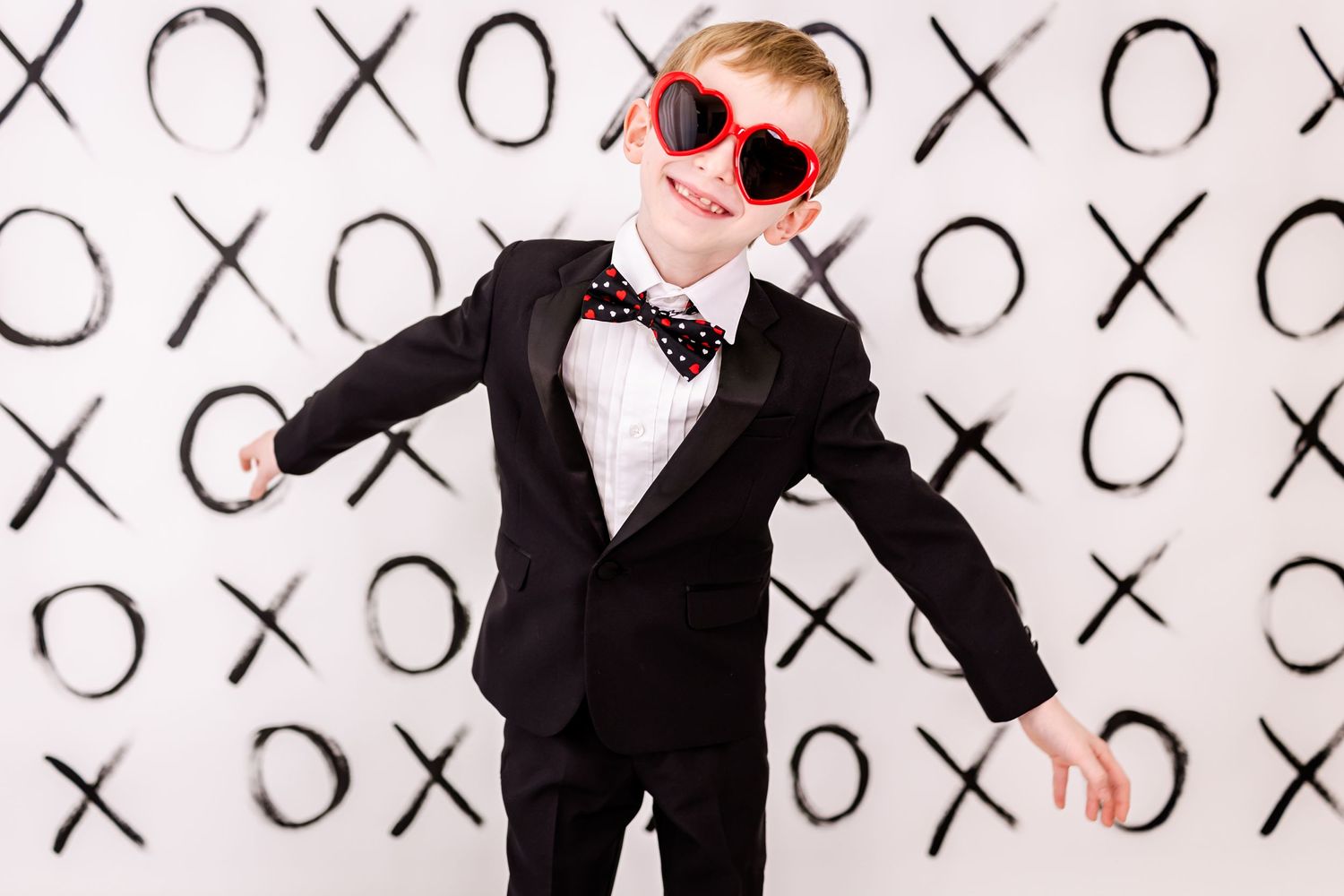 Someone in a black tuxedo and red heart-shaped sunglasses poses against a background with X's and O's pattern.