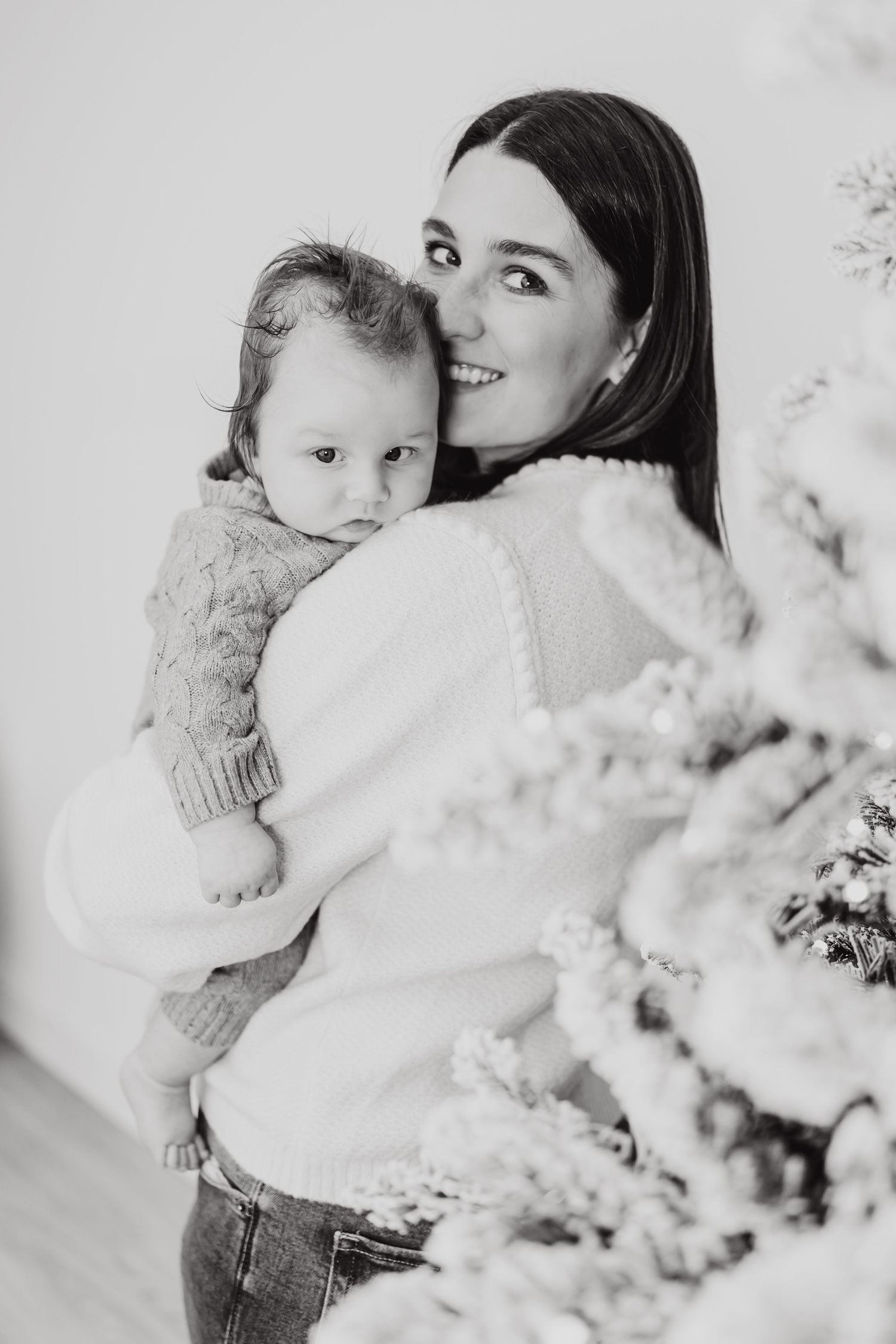 Black and white portrait of a mother holding a baby in winter clothing near a snow-covered tree.
