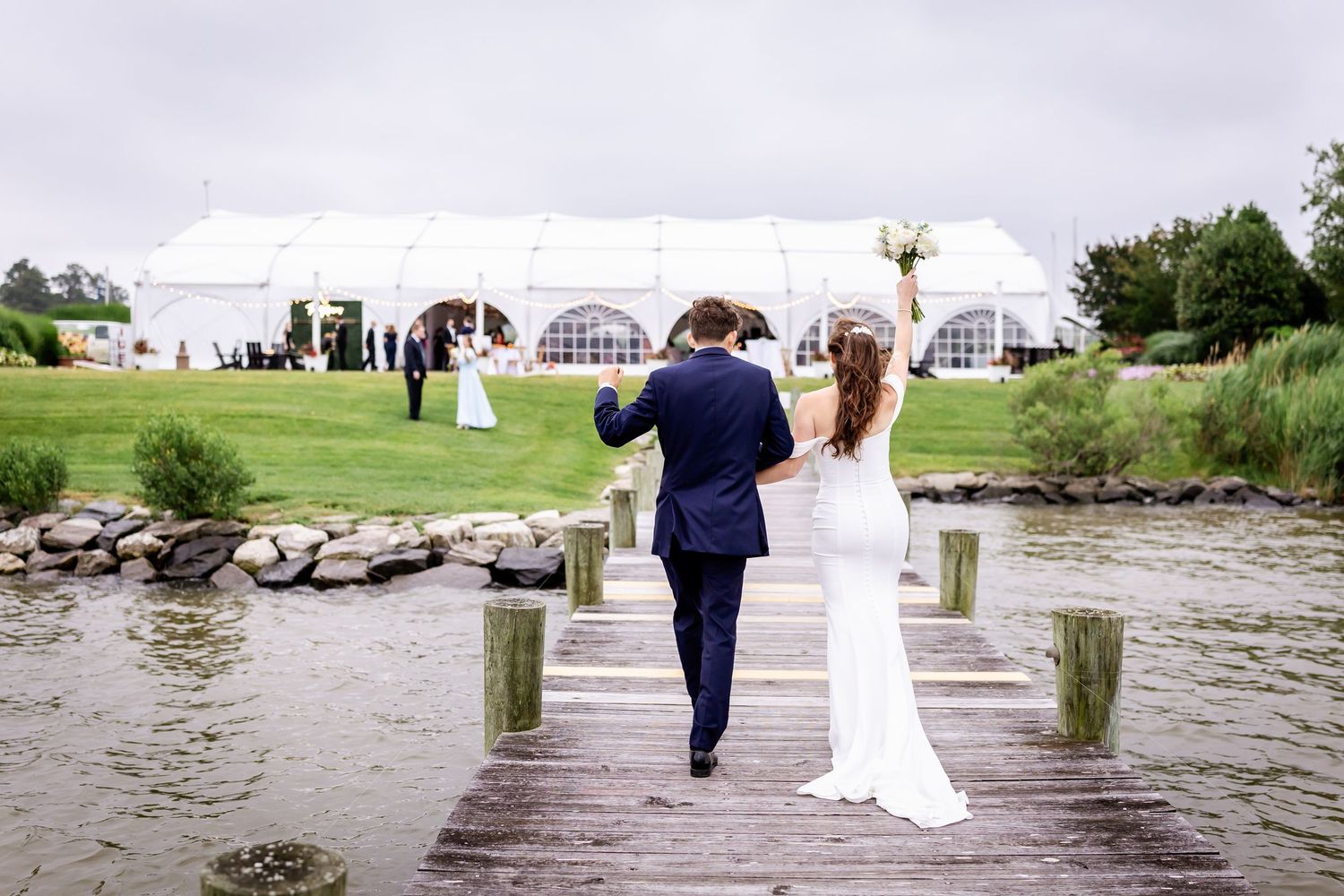A couple in wedding attire walks along a waterfront dock during a romantic seaside celebration.