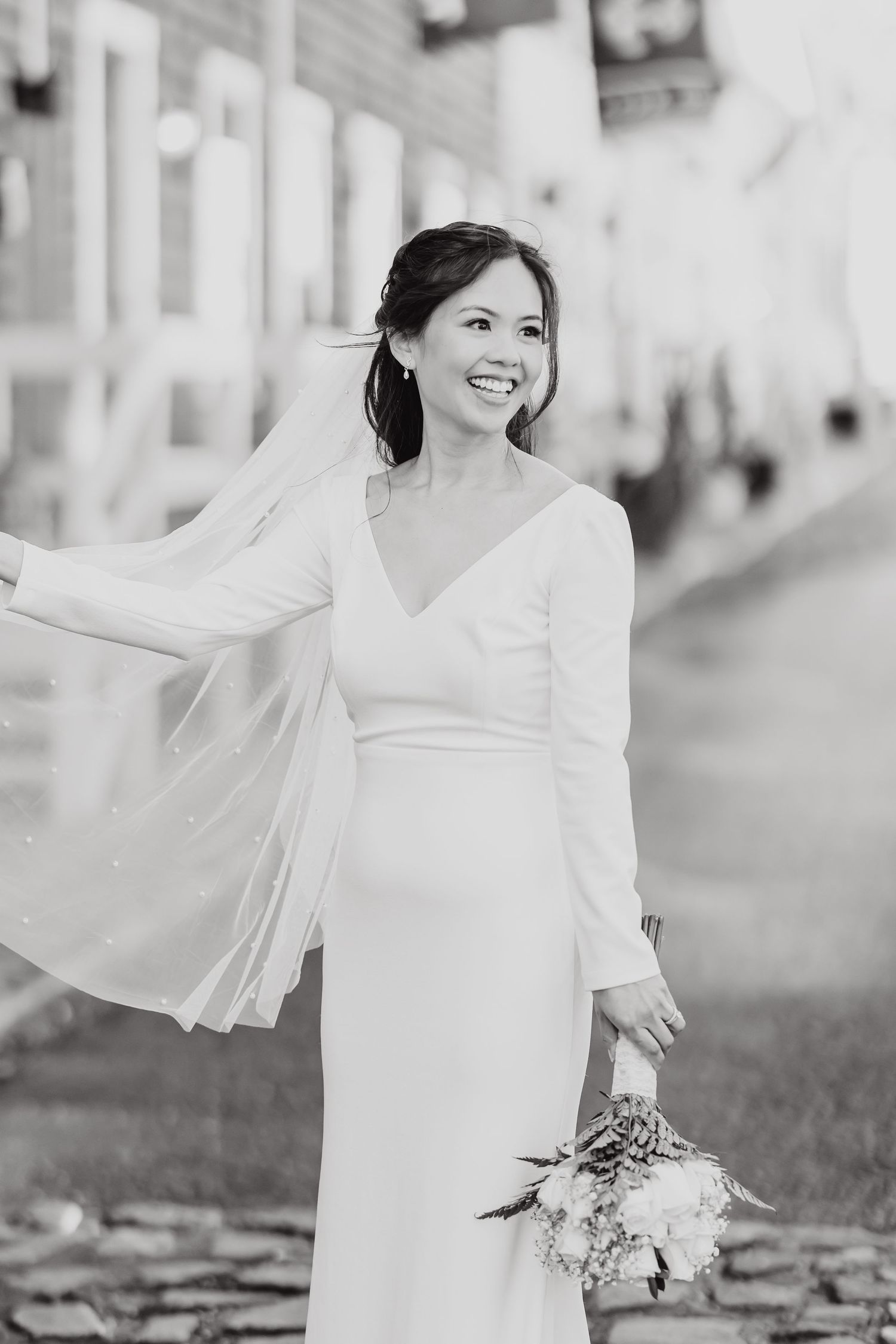 Joyful bride in simple white dress with flowing veil poses outdoors.