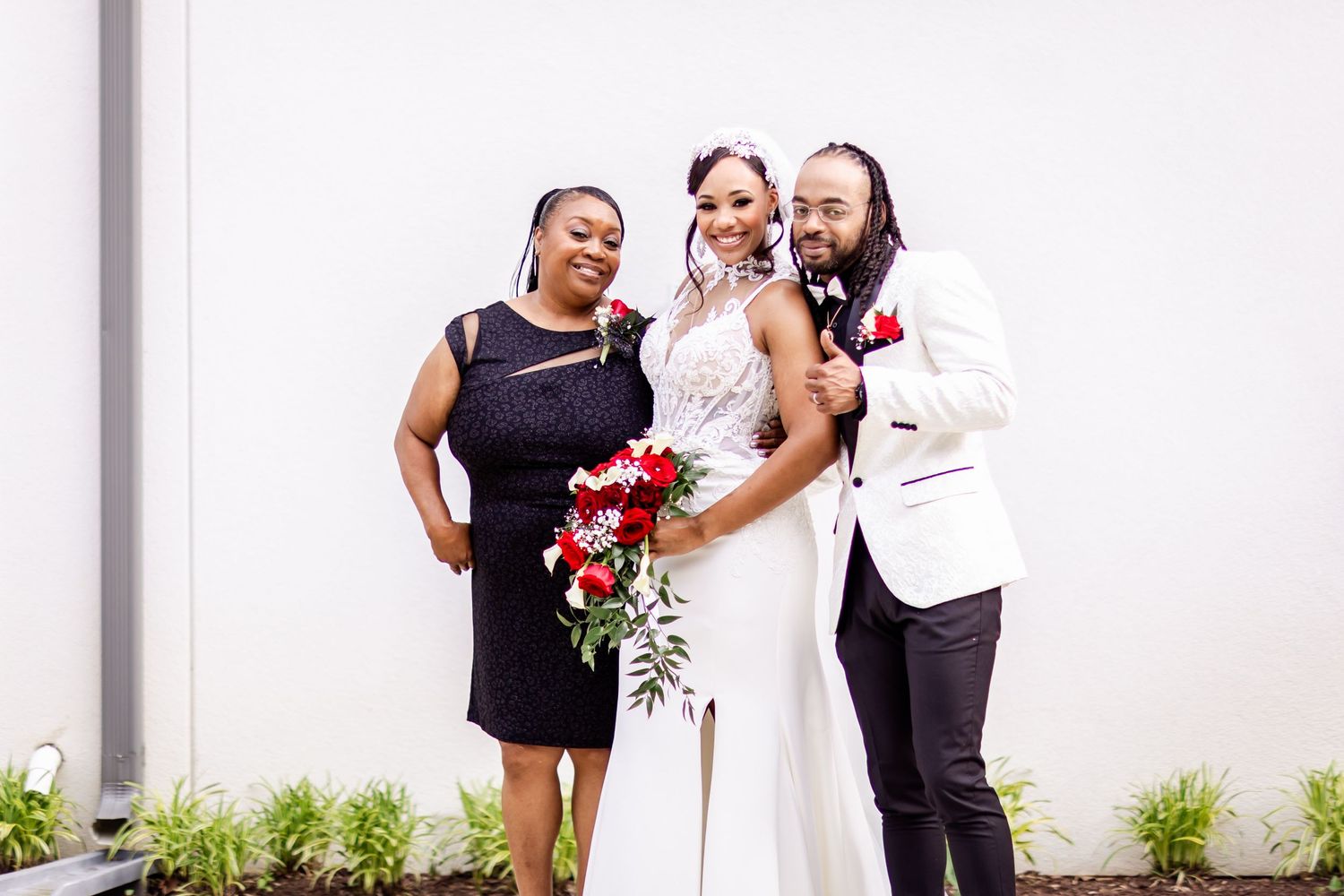 Wedding party members in black and white attire pose together with red floral bouquet against white wall.