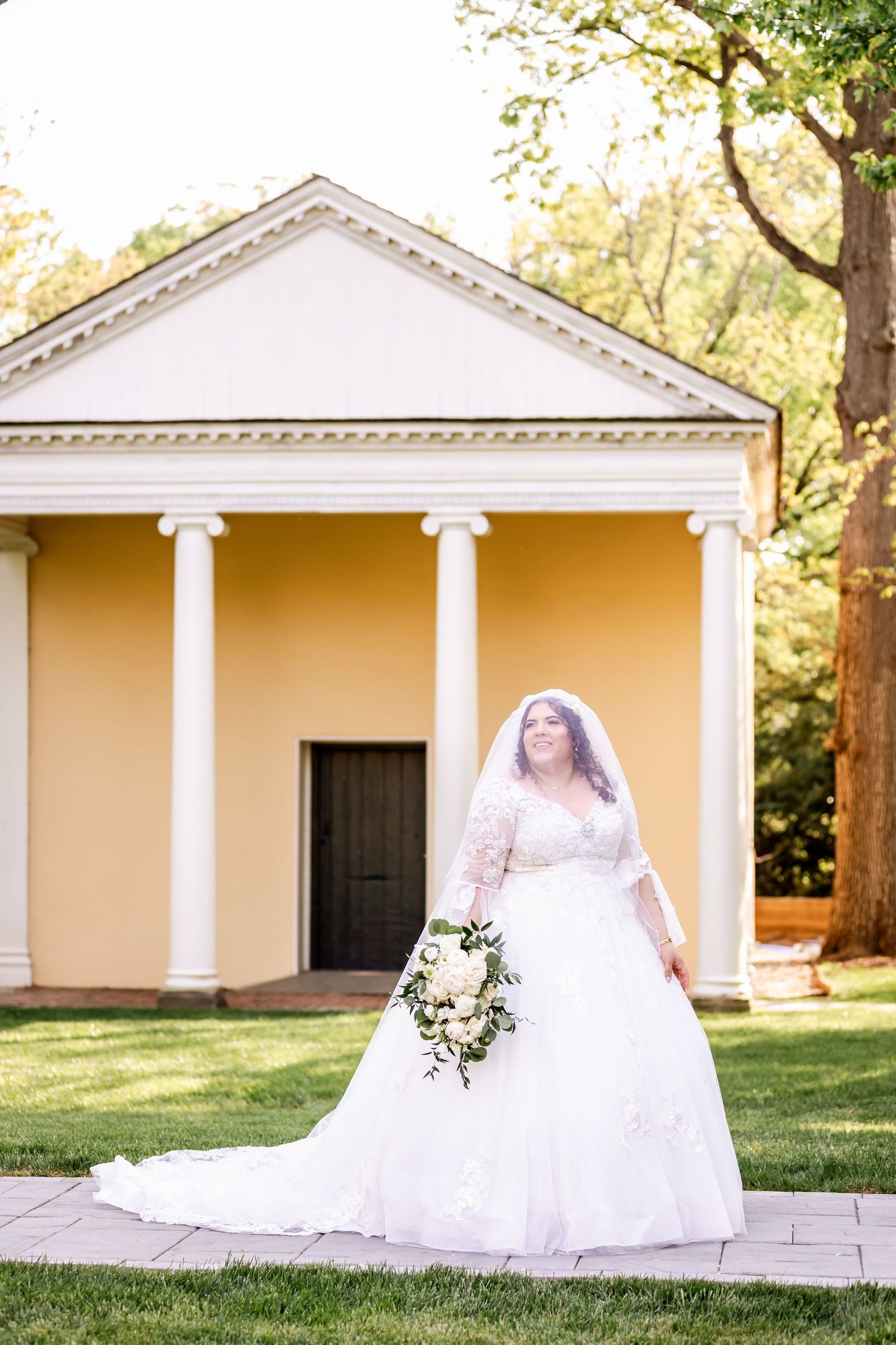Bride in flowing white gown poses with bouquet in front of classical yellow building.