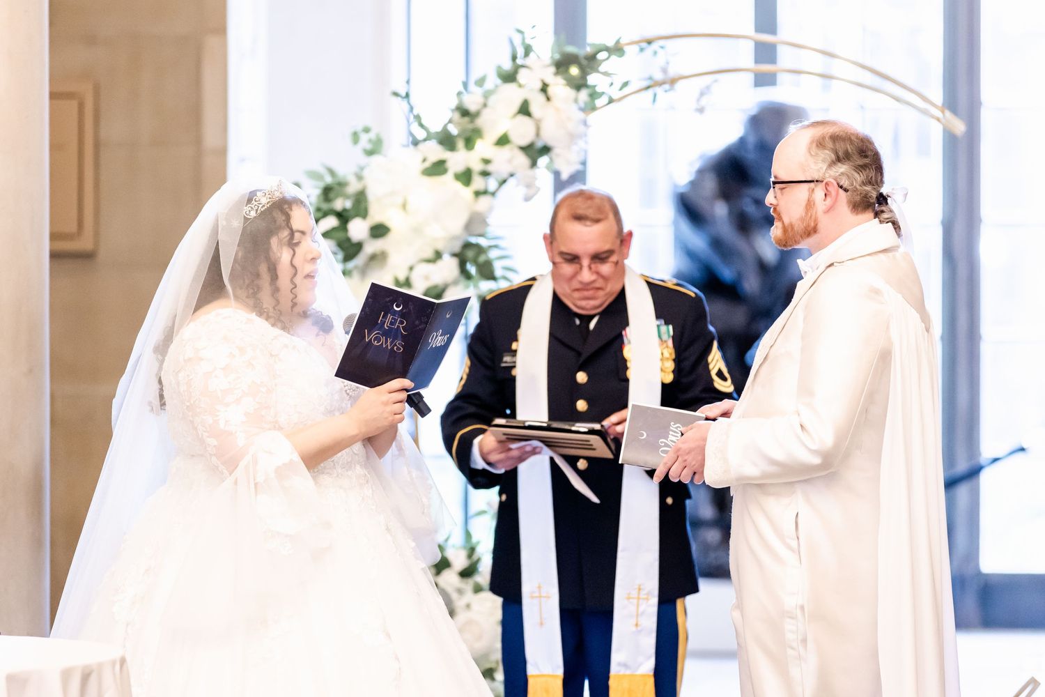 Wedding ceremony with couple and two officiants at the altar surrounded by white floral decorations.