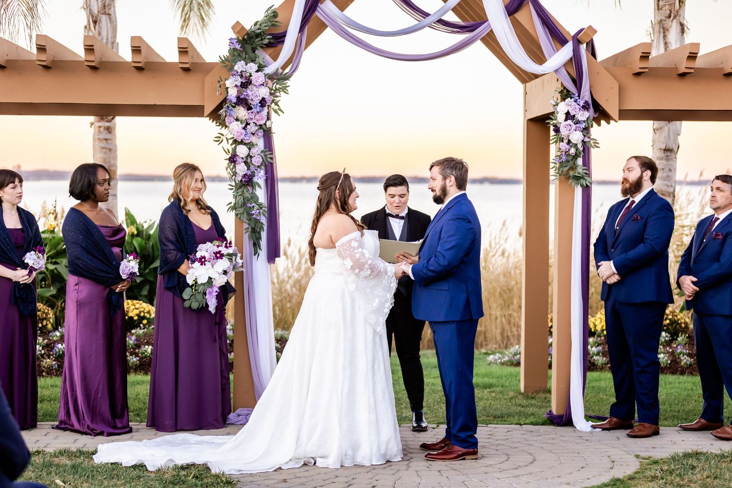 Wedding ceremony under decorated arch with purple draping and floral arrangements.