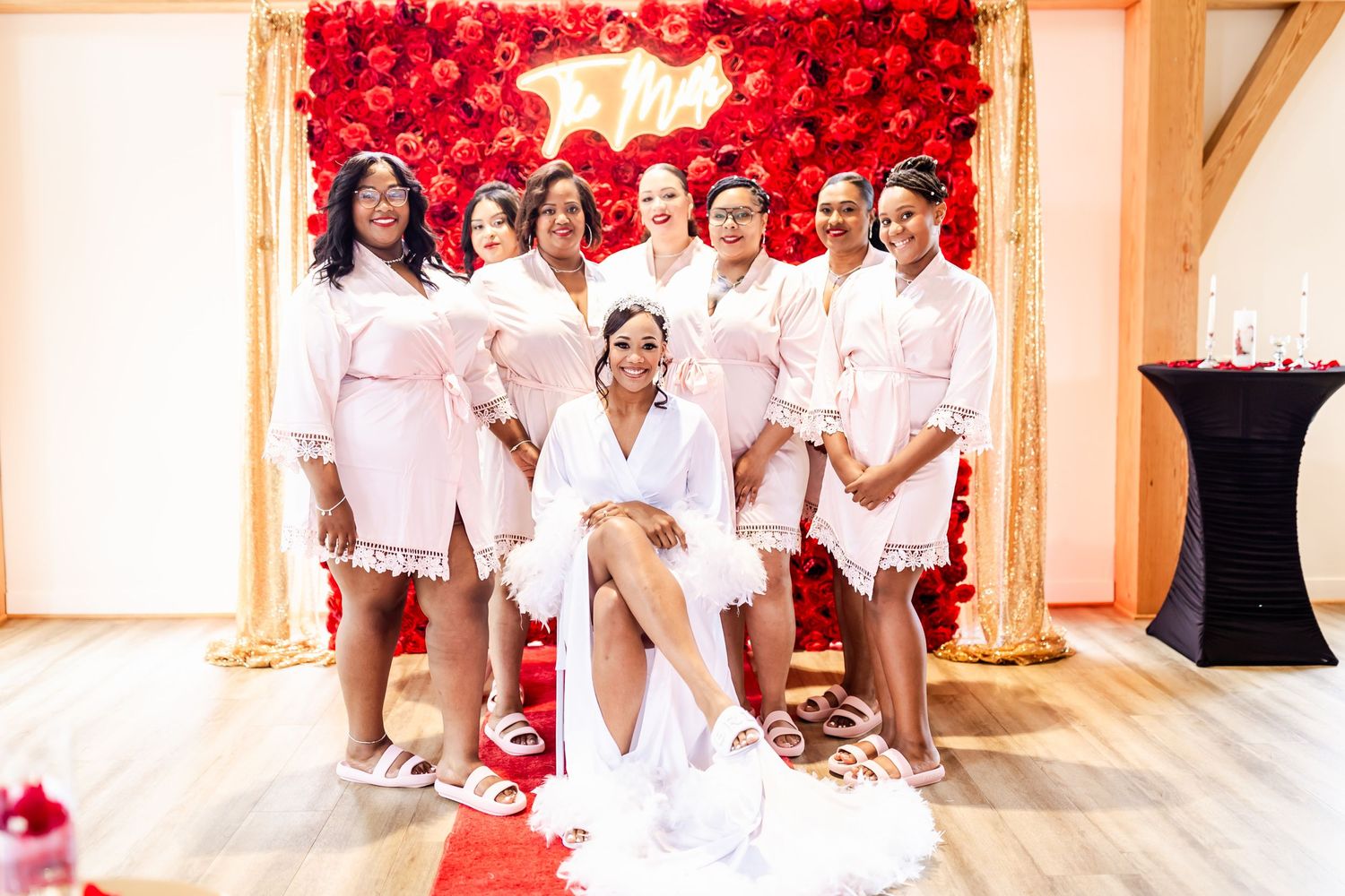 Bridal party posing in short white lace robes against red rose wall backdrop.