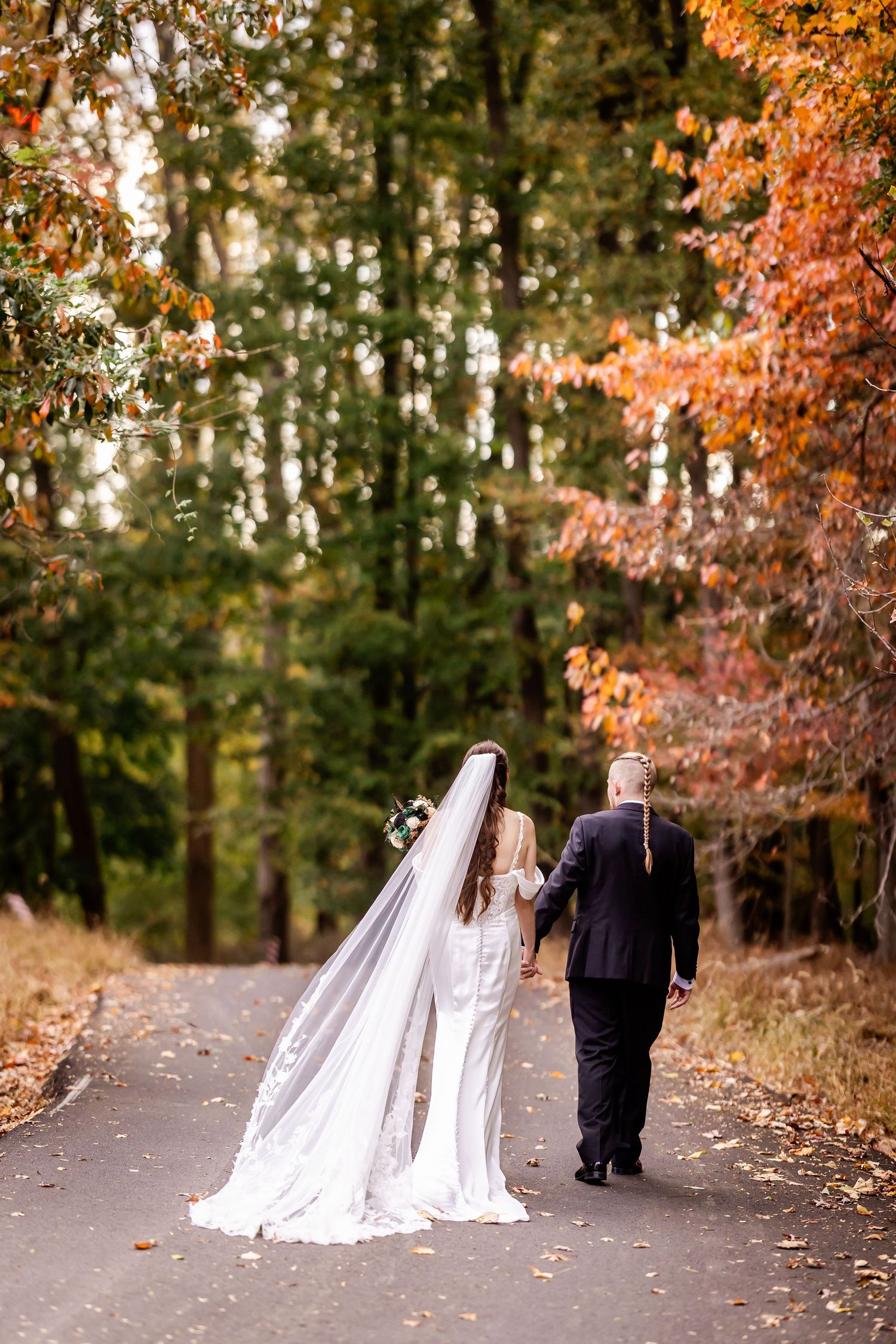 A couple walking down a tree-lined autumn path with colorful fall foliage overhead and a flowing white dress trailing behind.