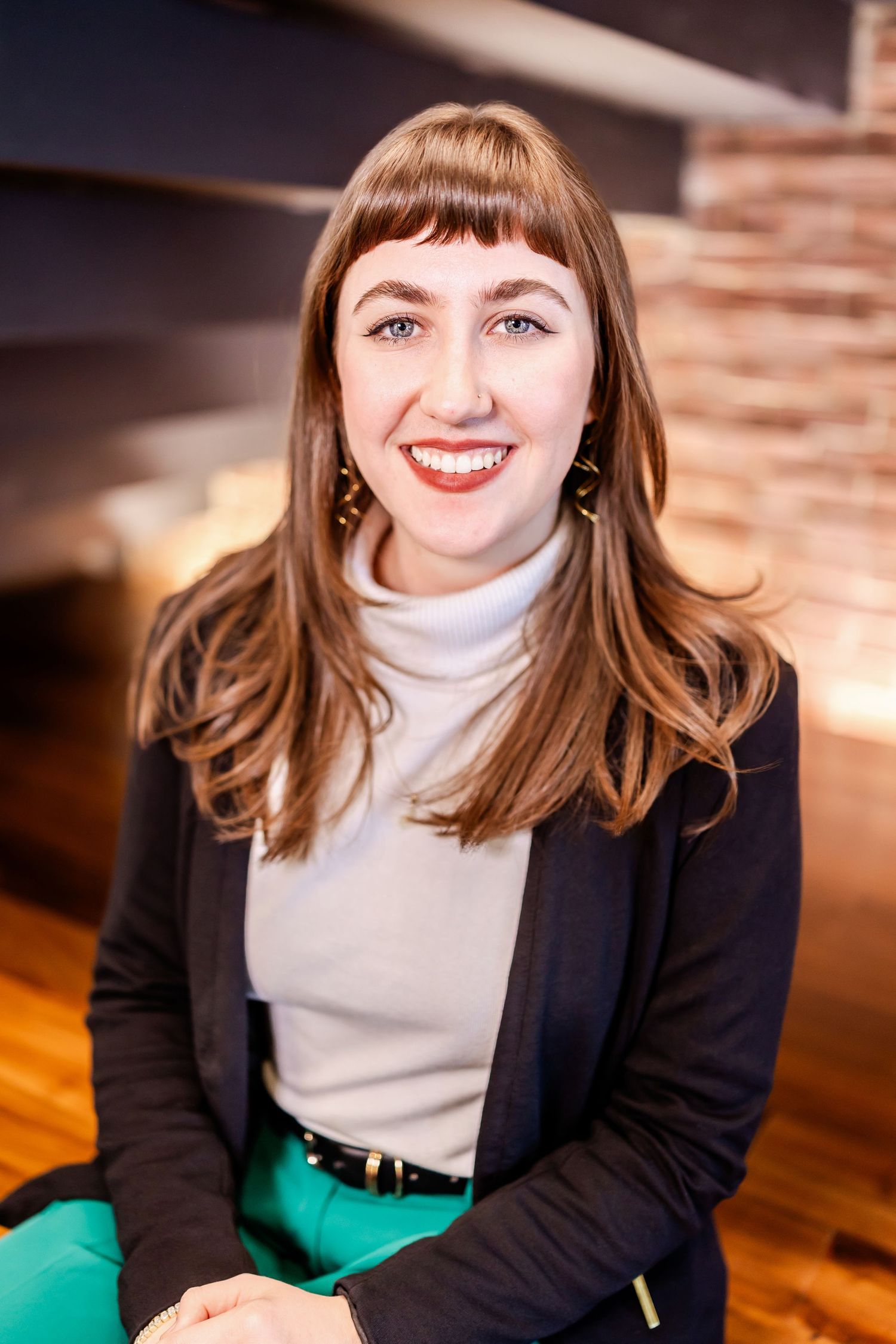Professional portrait of person in black blazer and turtleneck with bangs against wooden wall.