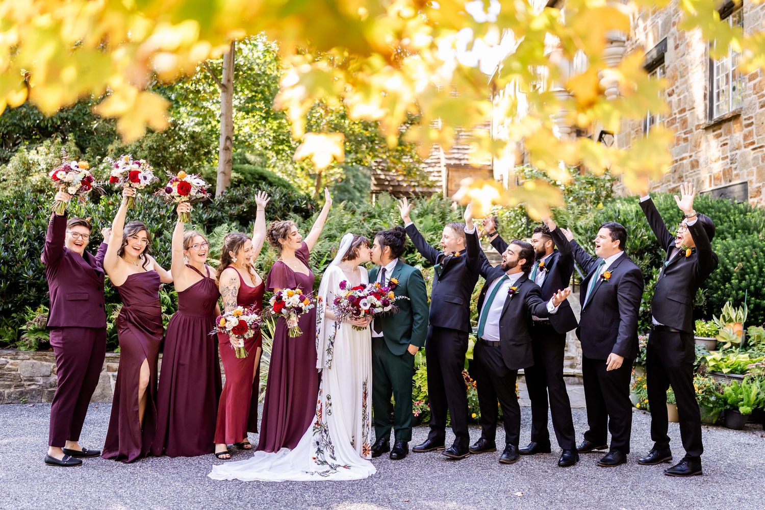 Wedding party poses outdoors in burgundy and black attire against a vibrant fall foliage backdrop.
