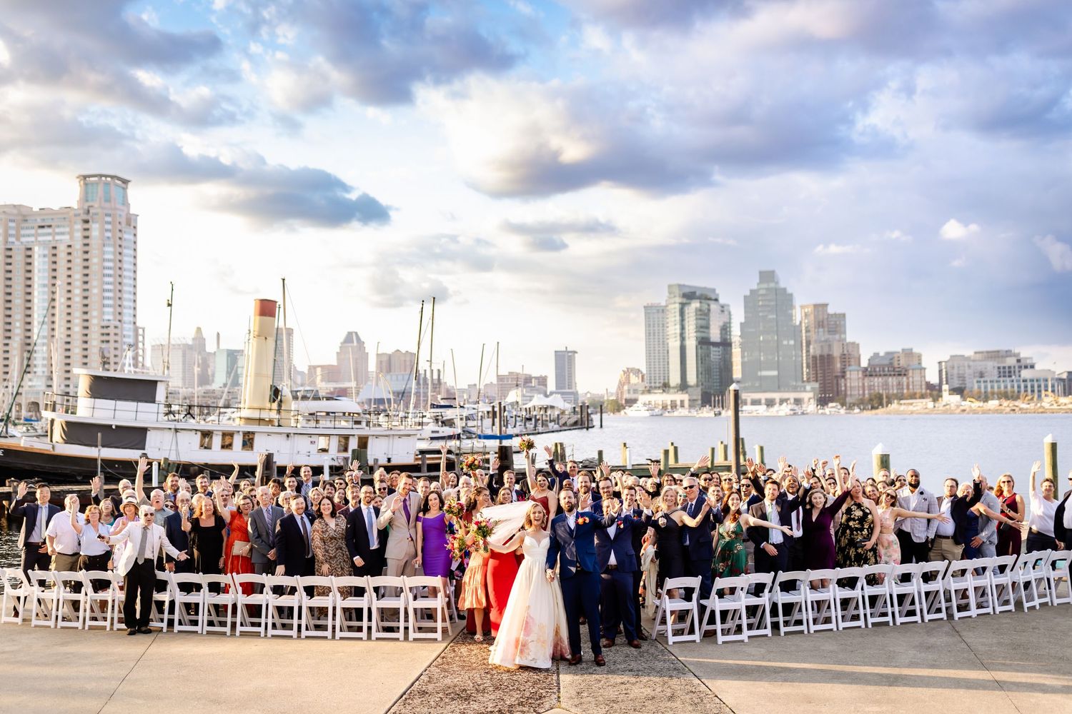 Panoramic view of wedding ceremony guests seated outdoors with city skyline and dramatic clouds in background.
