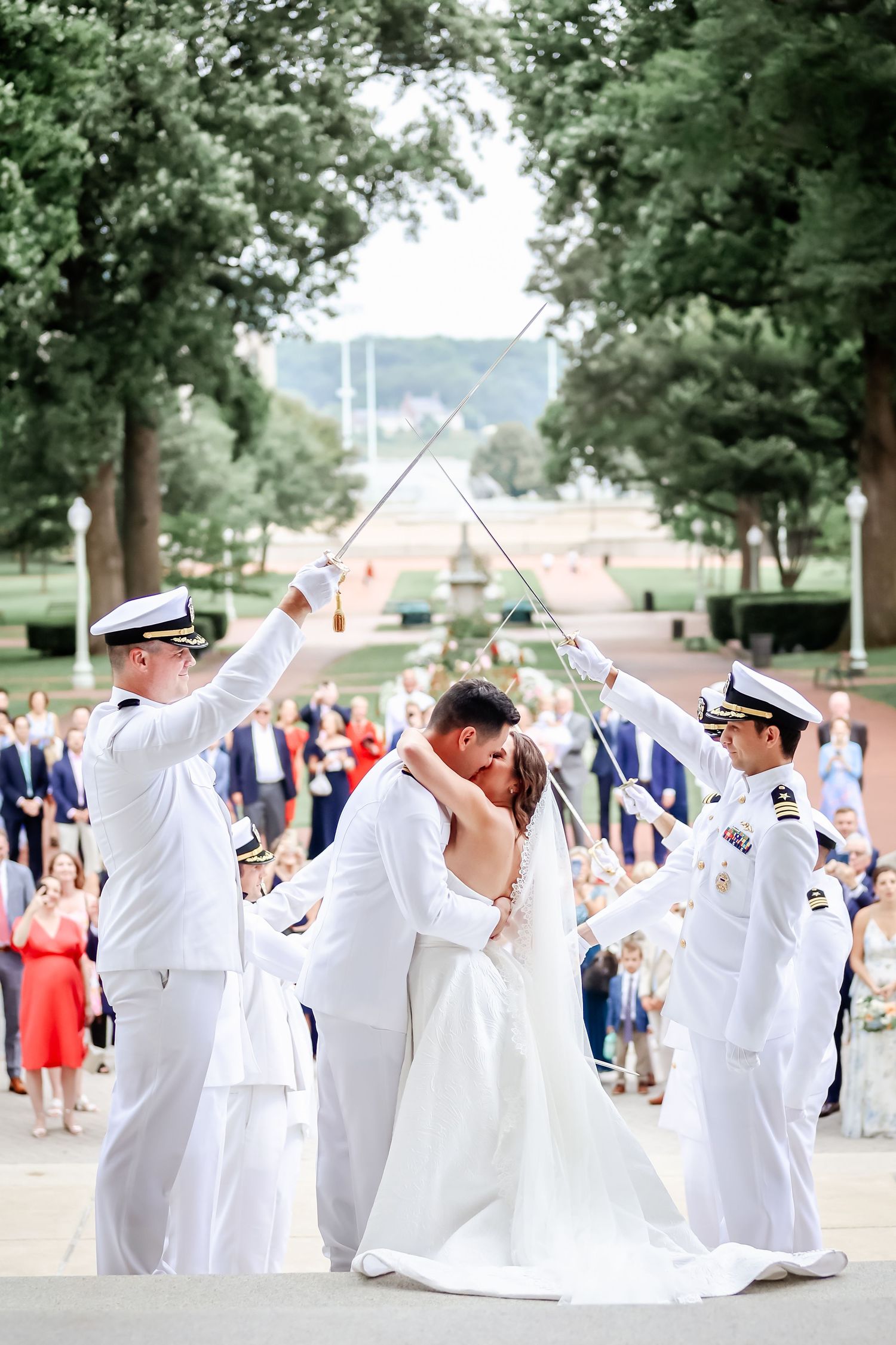 Wedding guests in white naval uniforms perform a synchronized group dance move at an outdoor celebration.