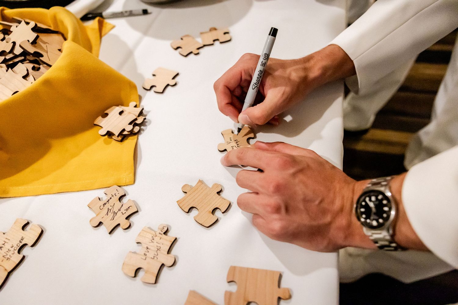 Hands signing wooden puzzle pieces on a yellow surface at a wedding event.
