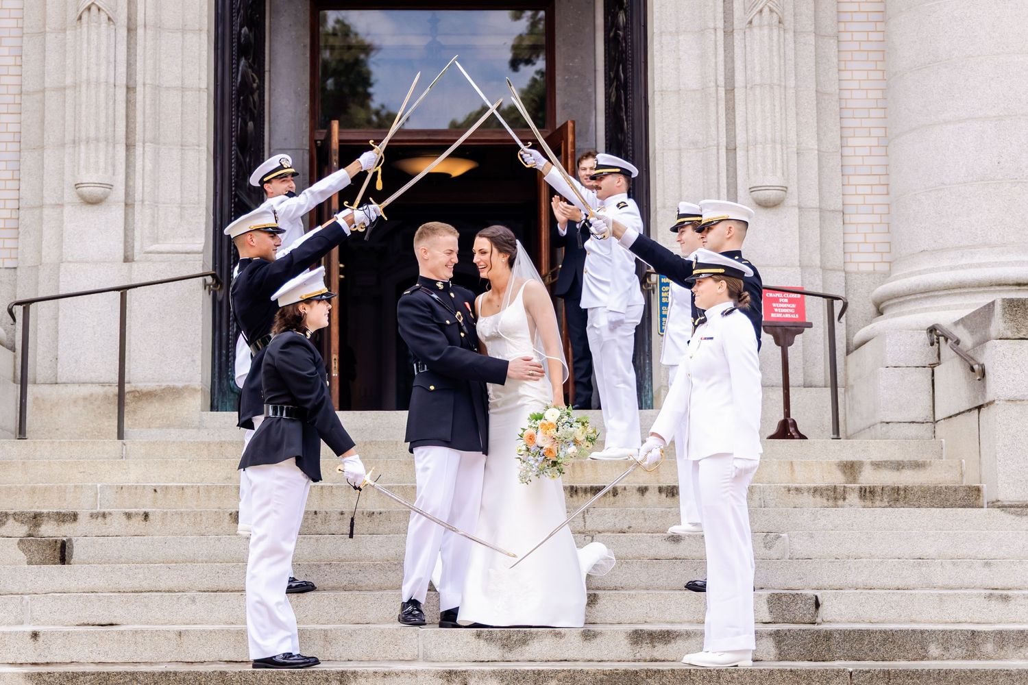 Military wedding party gathered on white stone steps of classical building.