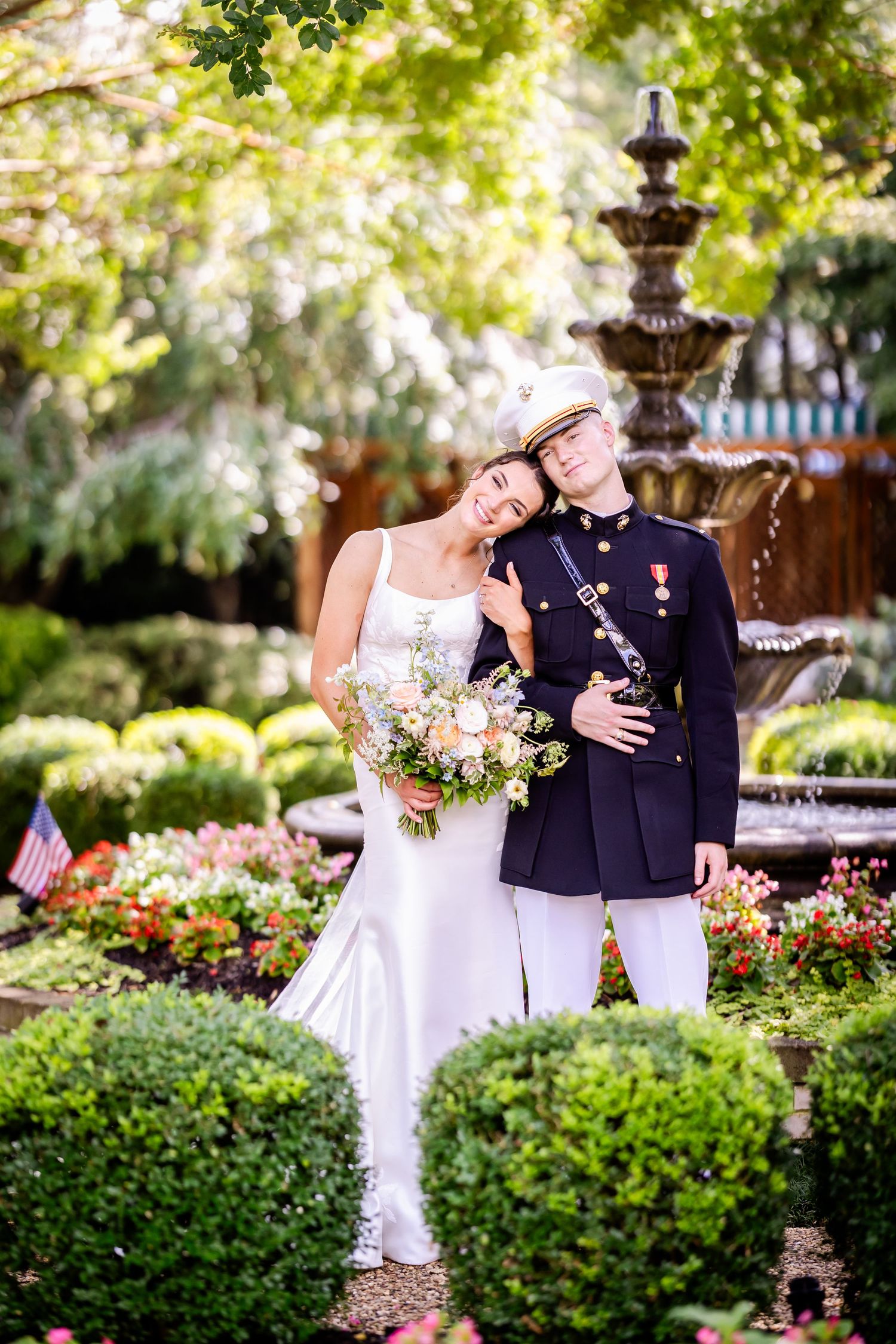 Military wedding couple poses in garden setting with fountain and lush greenery.