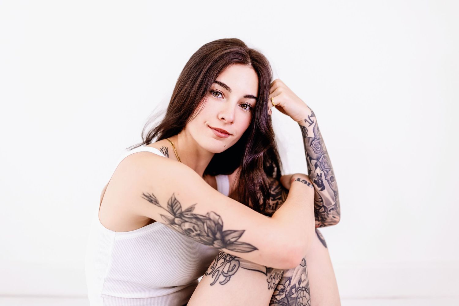 Woman with dark hair and tattoos poses in white tank top against light background.