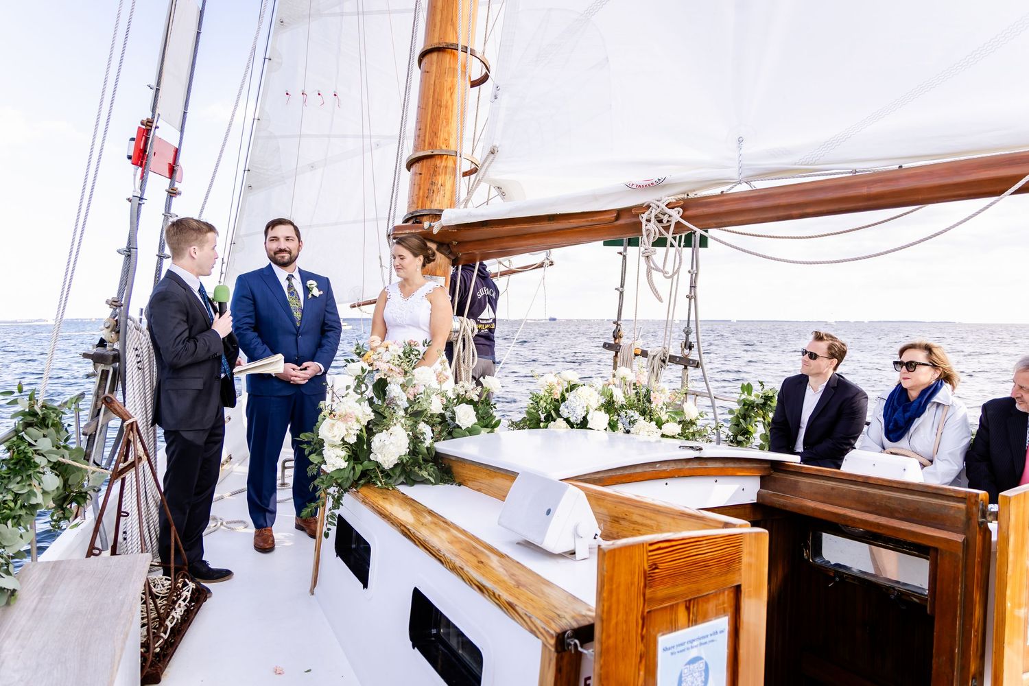 Couple exchanges vows during an outdoor wedding ceremony on a classic sailboat deck adorned with white floral arrangements.