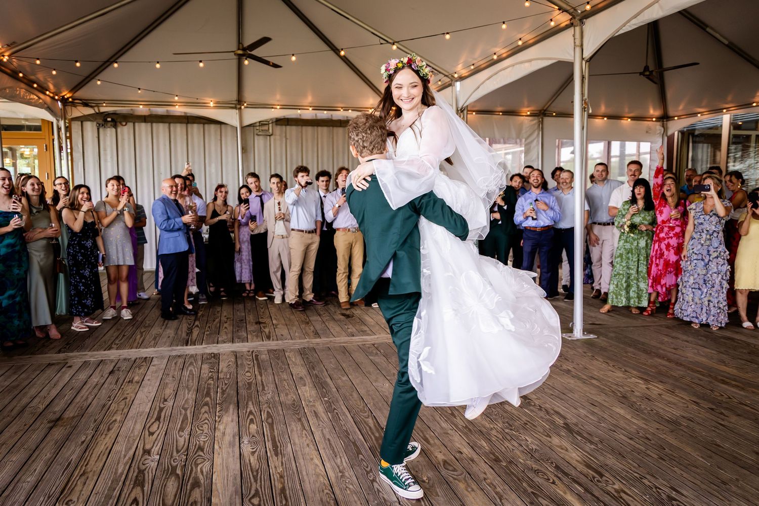 Wedding guests dance and celebrate on a rustic wooden deck under twinkling string lights at an outdoor reception.