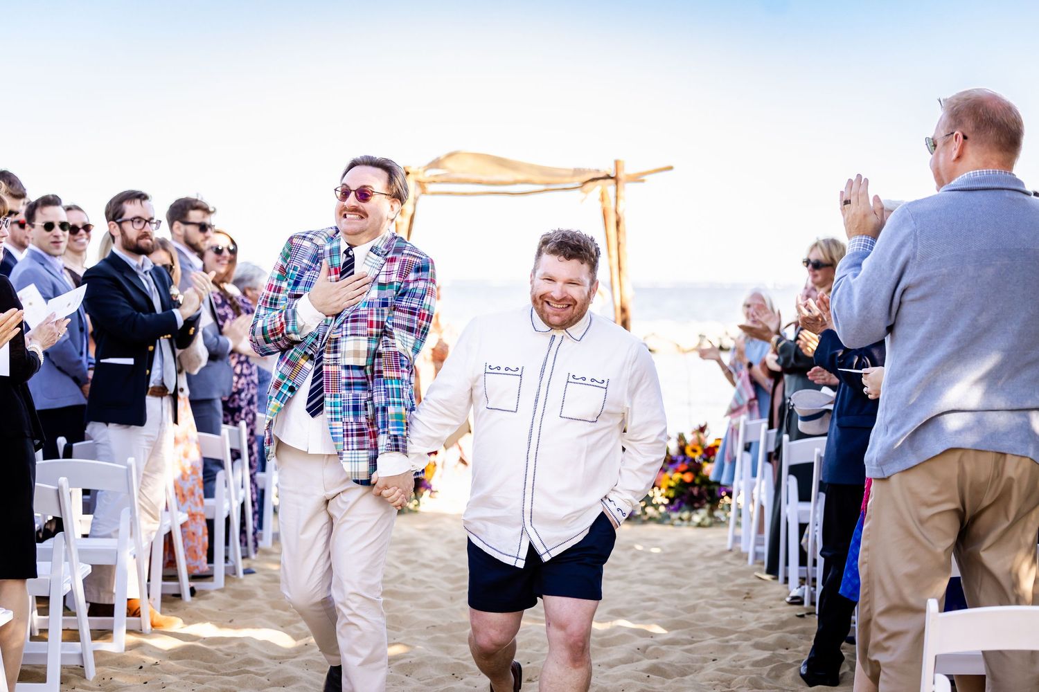 A group of people in formal attire gather at an outdoor beach wedding ceremony during sunset.