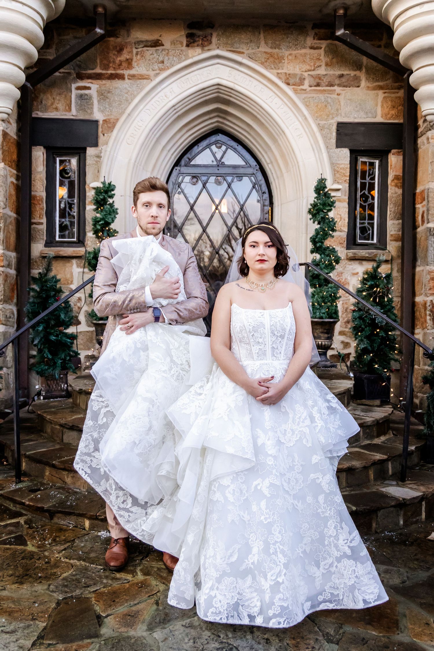 A wedding couple in white attire poses on stone steps in front of an ornate Gothic-style church entrance with arched doorway.