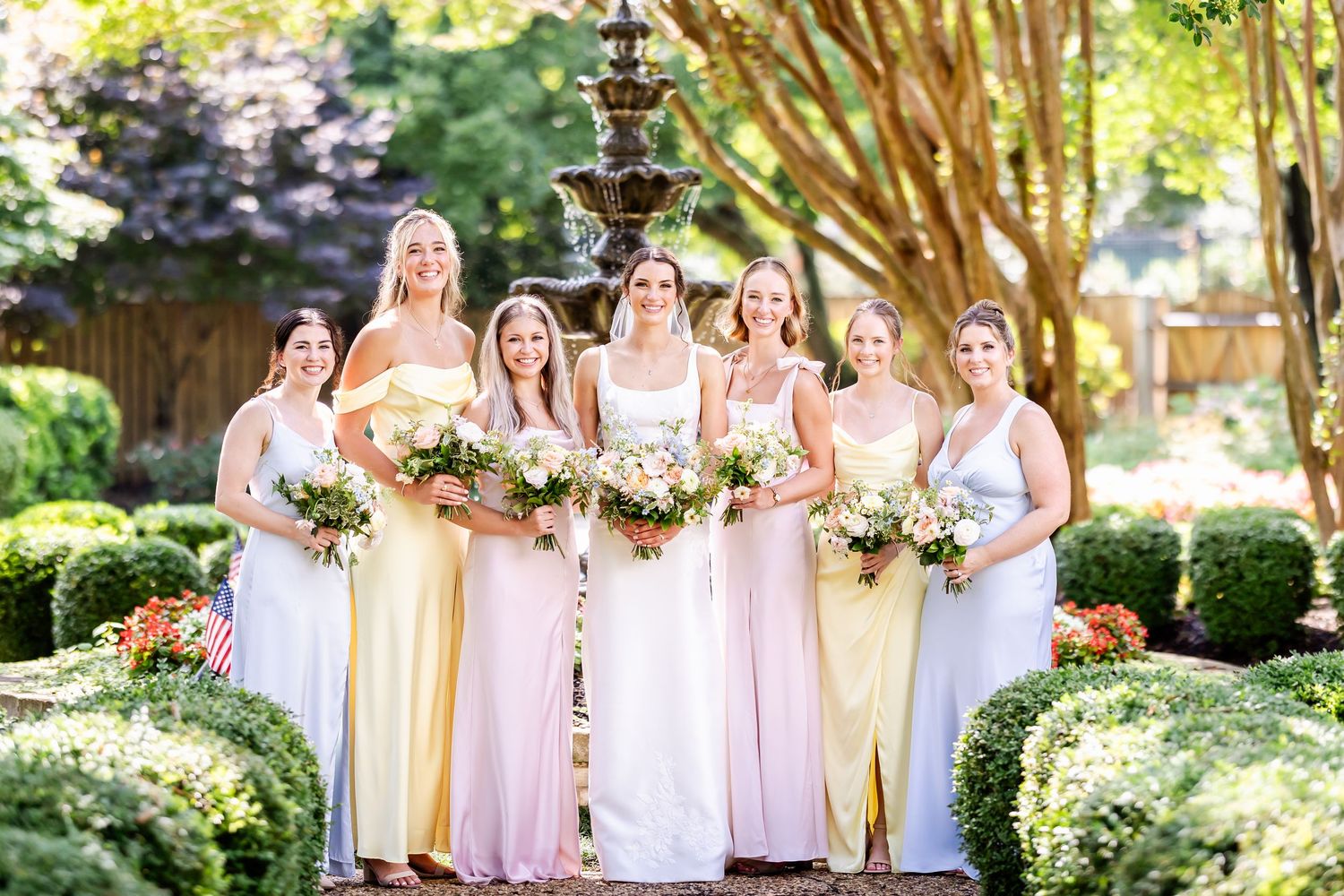 Bridesmaids in blush pink dresses pose together in garden setting with floral bouquets.