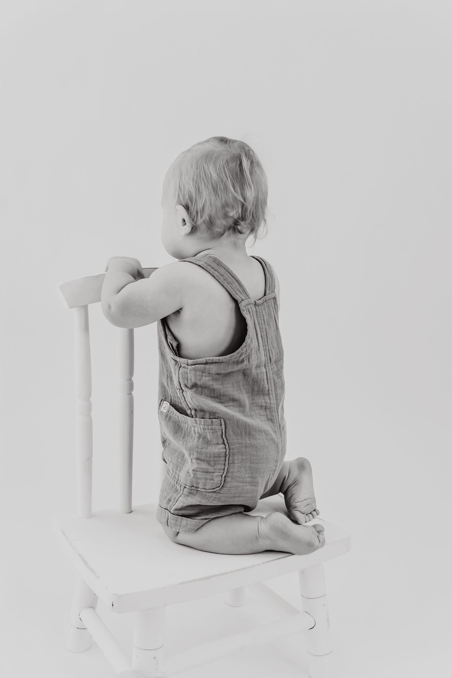 Baby in denim overalls sitting backwards on a white chair shown from multiple angles.