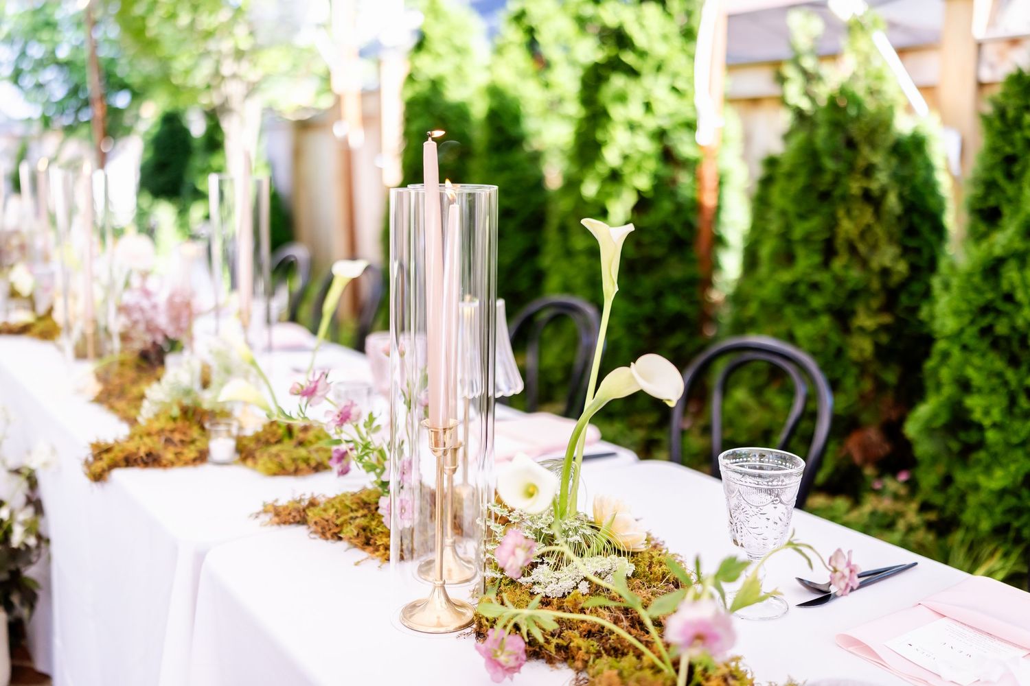 Elegant wedding table decor with glass candle holders, greenery and white linens in an outdoor garden setting.