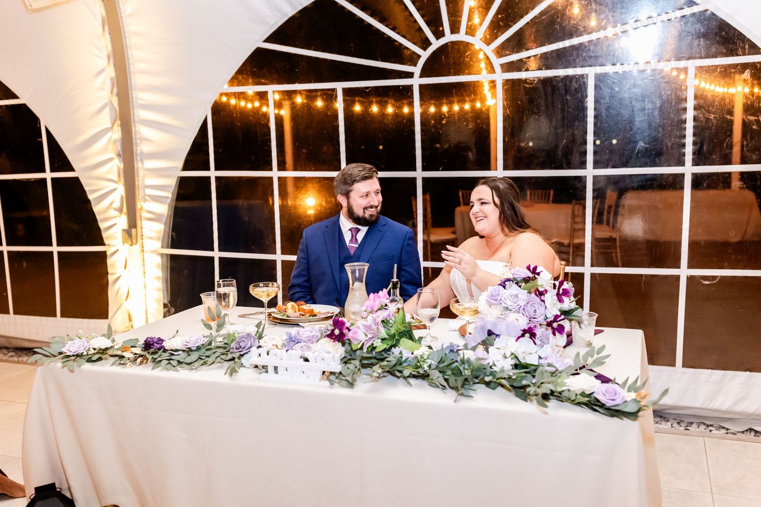 Newlyweds seated at reception head table decorated with greenery garland and white floral arrangements.
