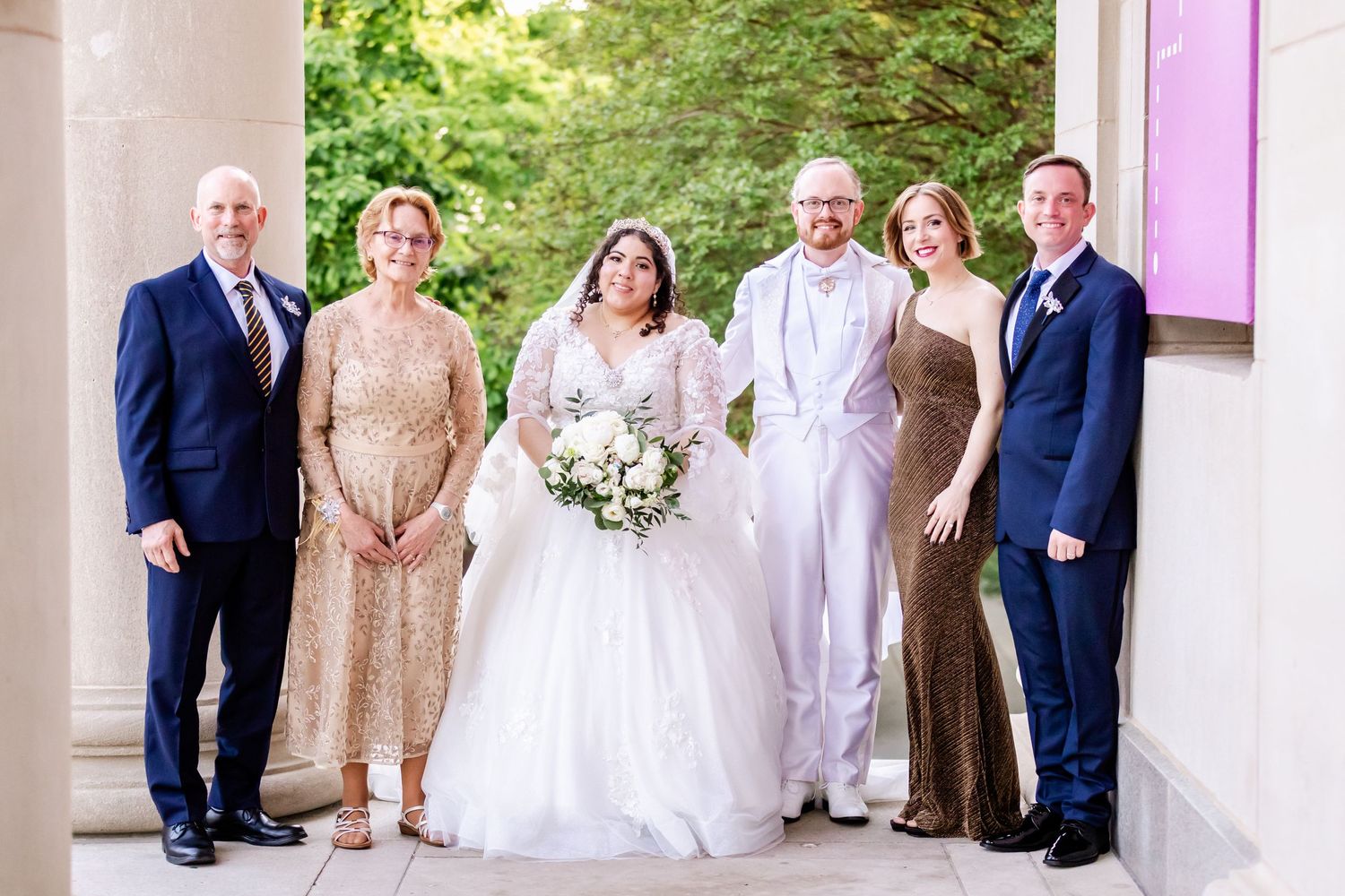 A wedding party of six people poses together against a light colored wall with greenery in the background.