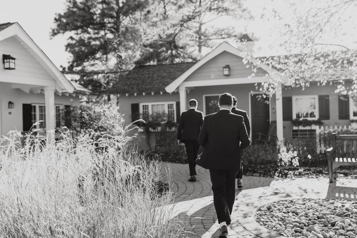 Black and white photo of figures walking along residential sidewalk with quaint cottage-style houses in background.