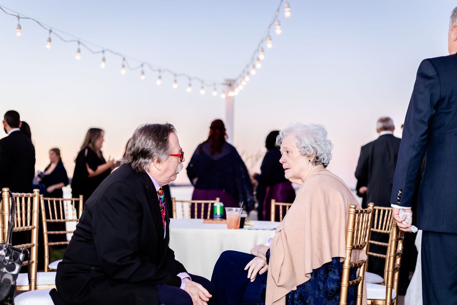 Wedding reception guests seated at tables under string lights during twilight hours.